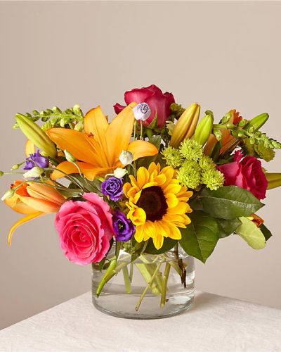 <p>This bouquet features sunny sunflowers, hot pink roses, purple double lisianthus, orange LA Hybrid lilies, yellow snapdragons, green button poms, and fresh greenery. Arranged in a clear glass vase, the flowers take center stage, letting their natural brilliance shine through with effortless charm.</p>
