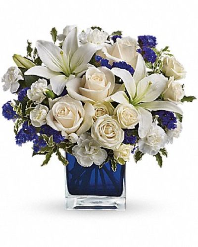 <p>Luxurious crème roses and pure white lilies paint a peaceful picture inside a blue cube. Crème roses, white asiatic lilies and white miniature carnations are mixed with bursts of purple statice and green pitta negra.</p>